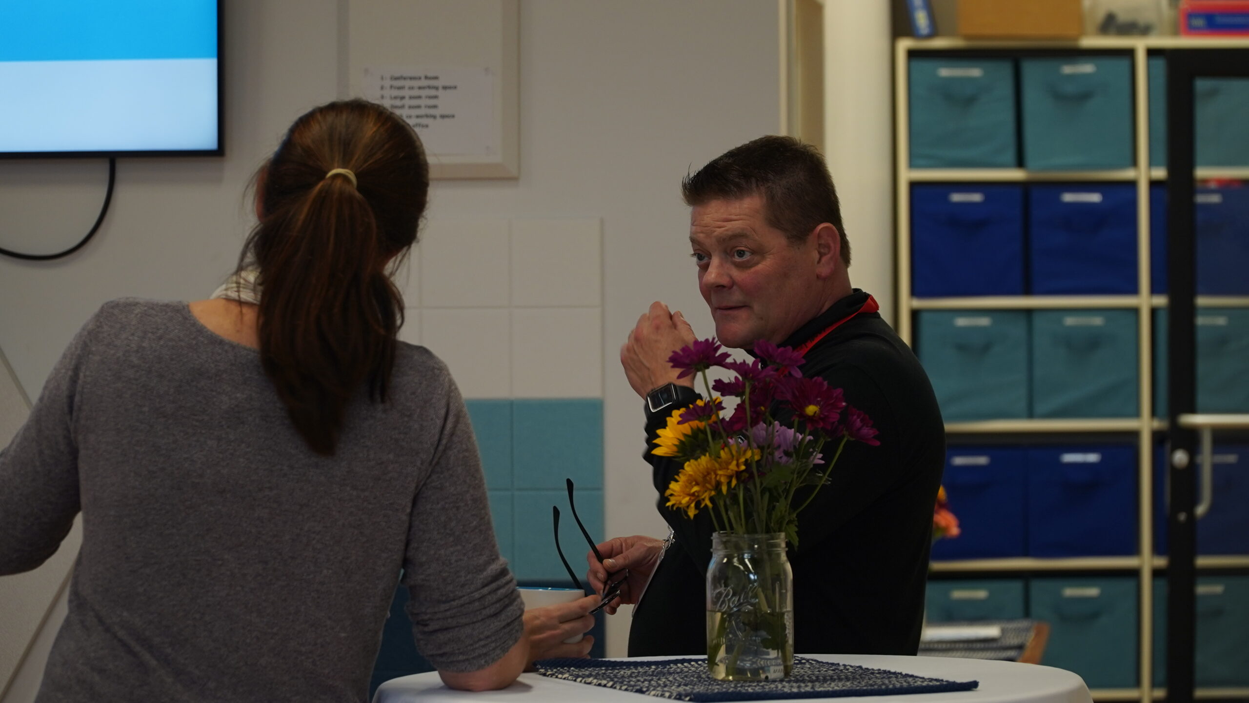 Two event attendees converse with a mixed bouquet of flowers on a high table in the foreground.