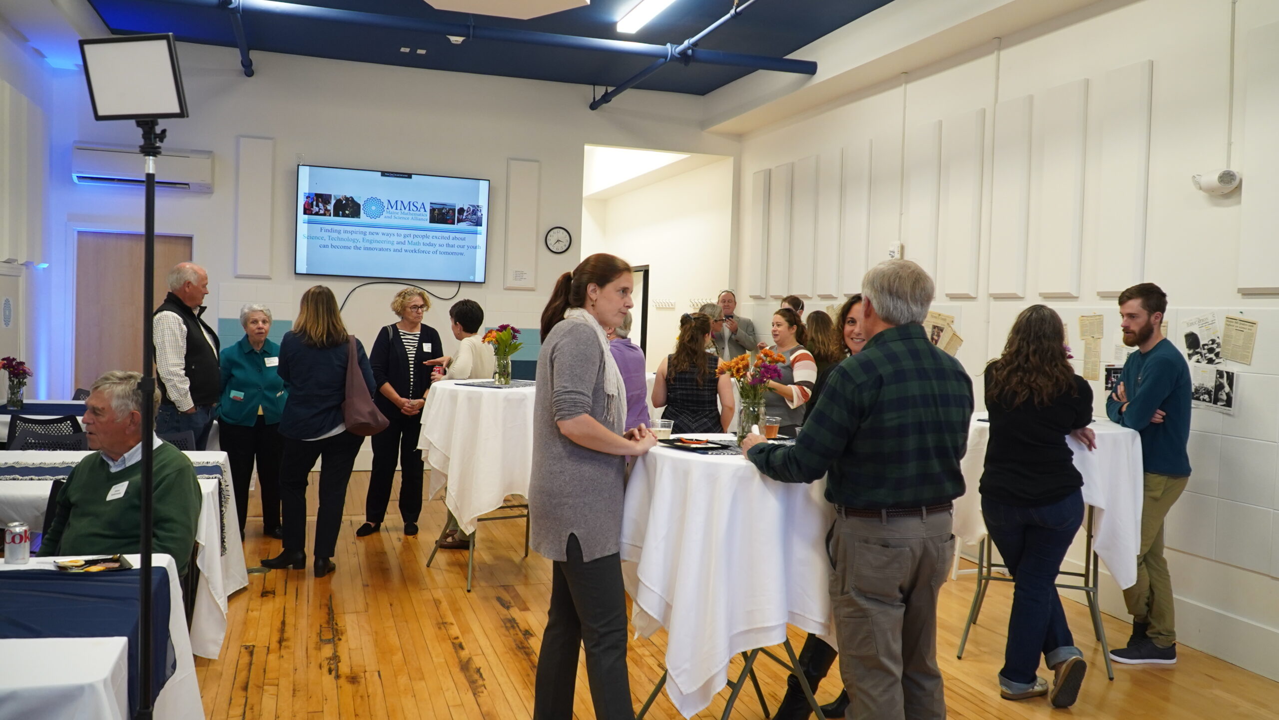 A group of attendees converse and a powerpoint slide showing a current project can be seen behind them.