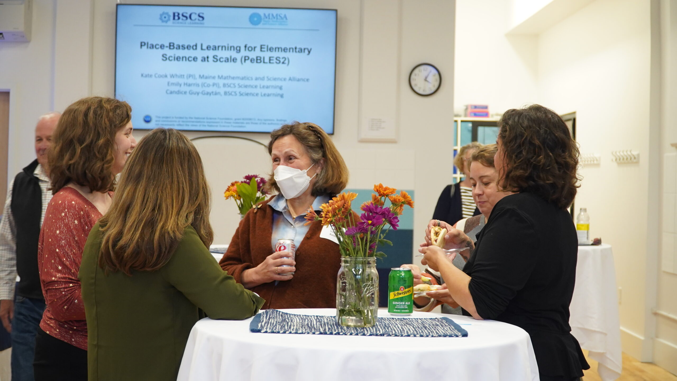 Multiple attendees converse and a screen in the background shows a slide describing the pebbles project.