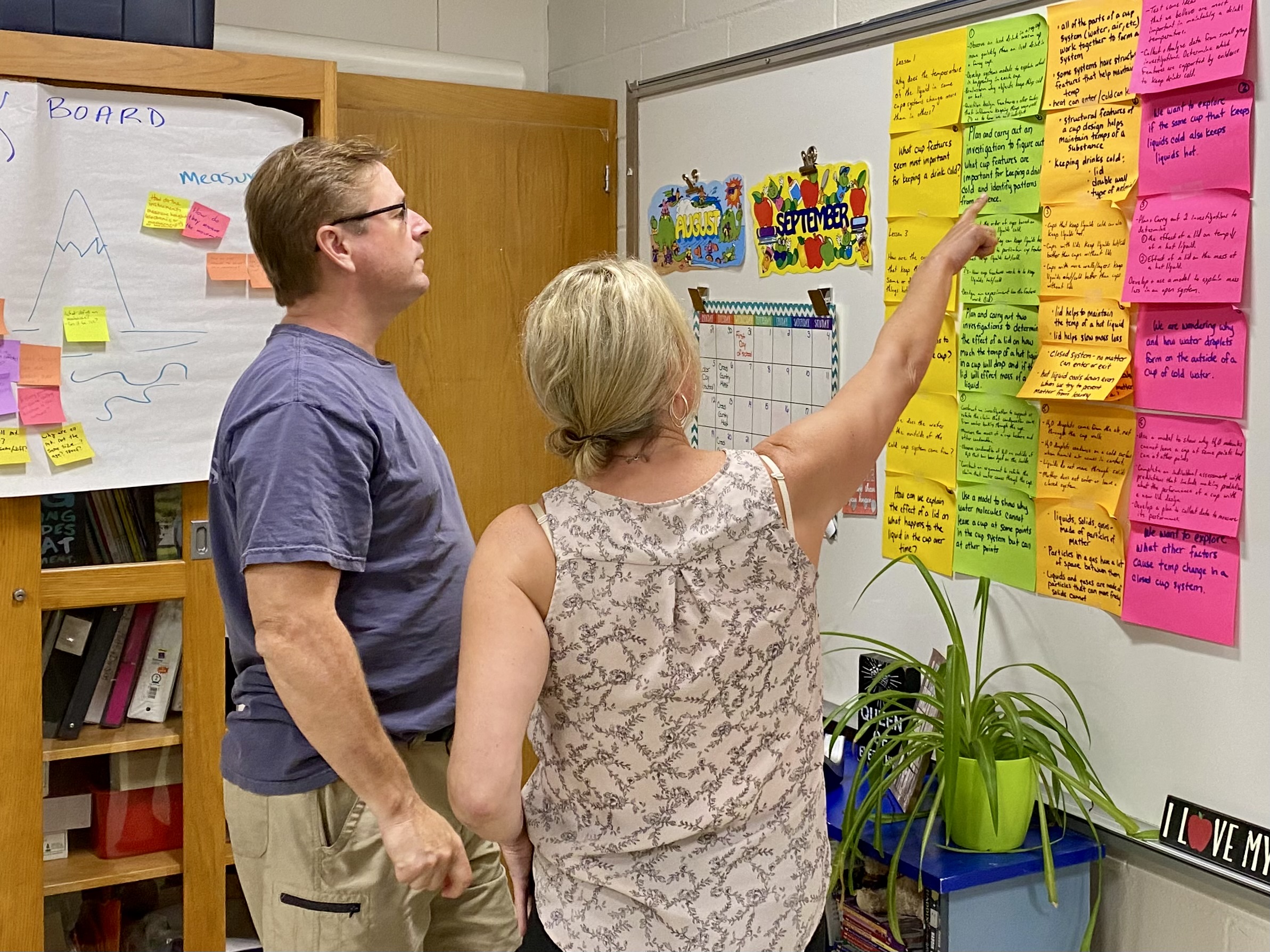 Two teachers review a board with sticky notes organized in columns.