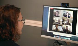 Photo of woman looking at a webcam on top of a screen showing a Zoom session with a group of participants.