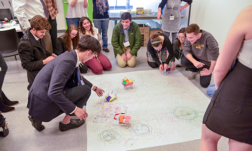 Students crouch in a circle to look at a small robot that draws on a large sheet of paper when it moves.