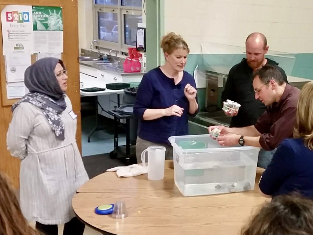 Adults converse around a table with a box of materials on it in a schoolroom.