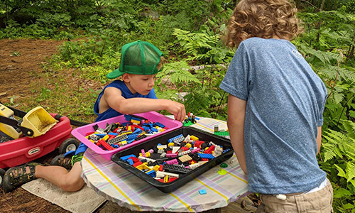 Two kids play with legos on a table outdoors surrounded by greenery.