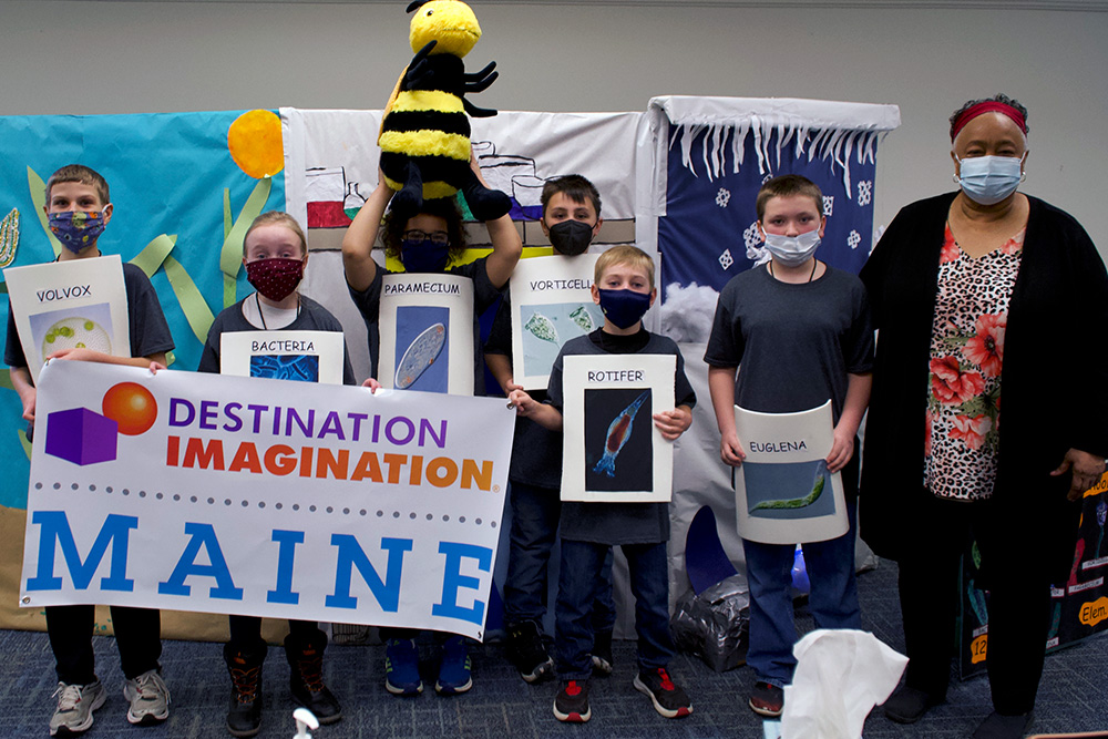 A teacher is shown with students holding pieces of paper with images that match titles situated above them, which read: "volvox," "bacteria," "paramecium," "vorticella," "rotifer," "euglena." They are also holding a "Destination Imagination Maine" sign and a stuffed bumblebee.