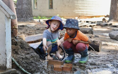 Two children wearing bucket hats crouch down outside next to a puddle. Both children stare intently at a stream of water one child is positioning over the mud using a garden hose.