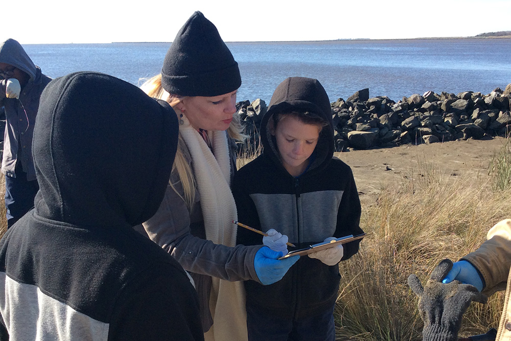 A photo of students and a teacher in cold weather clothing standing by the ocean looking at a clipboard together.
