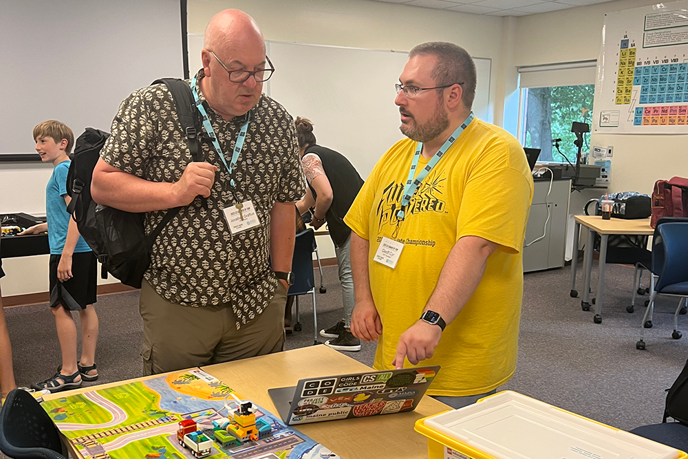 Two educators converse in front of a computer while wearing lanyards for a conference.