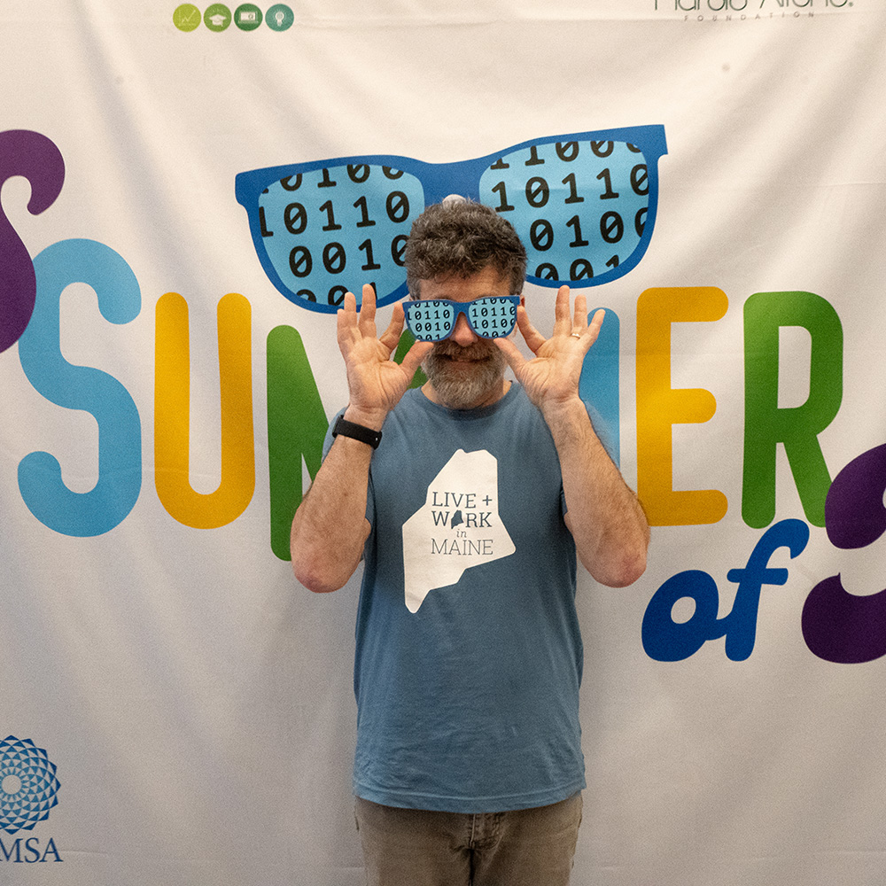 An educator stands smiling and wearing glasses printed with binary code on the lenses in front of a banner with the CS Summer of Fun logo on it.