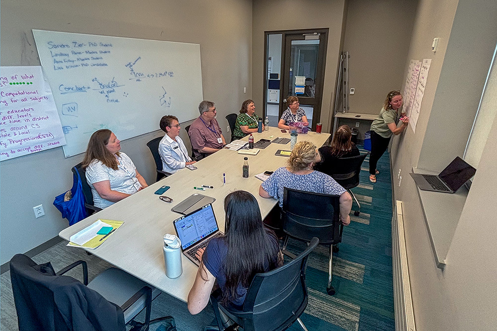 A group of people converse in a conference room with one person taking notes on a poster-sized piece of paper.