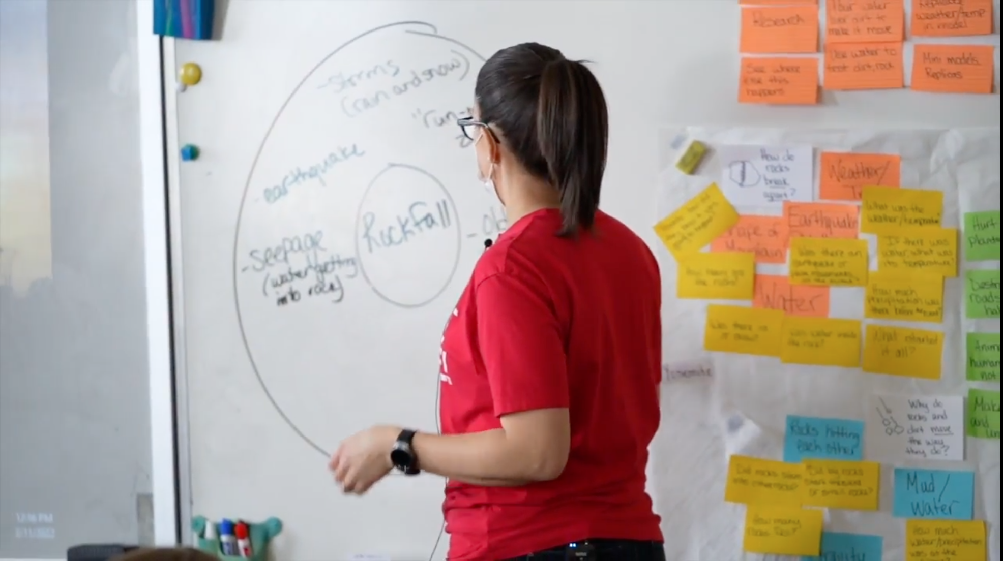 A teacher stands back to the camera in front of a whiteboard with a drawing of a chart that shows the words "Rockfall" and "earthquake" visible; to the right there is a collection of sticky notes mounted on a poster-sized paper mounted on the whiteboard.