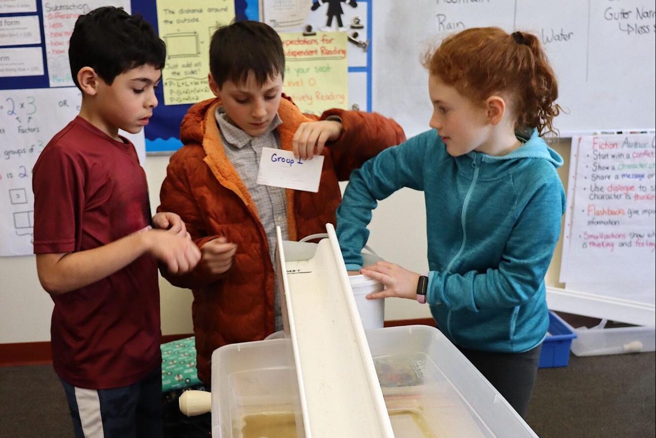 Three children stand next to a bin with sand, water, and an inclined plastic chute.