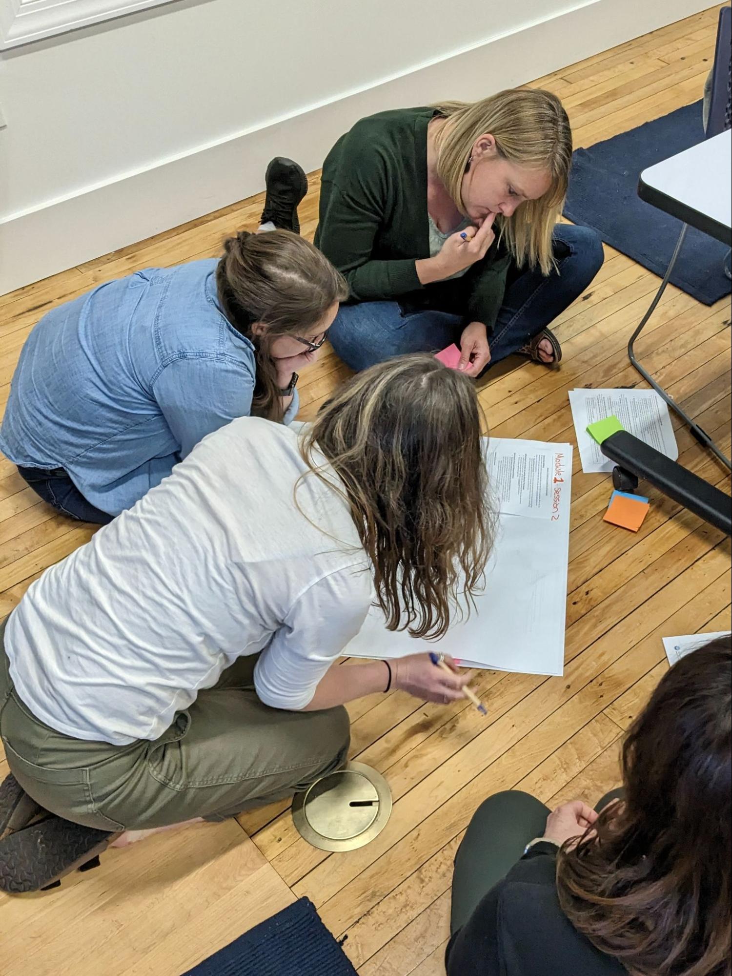 Three individuals collaborate on a piece of paper on the floor.