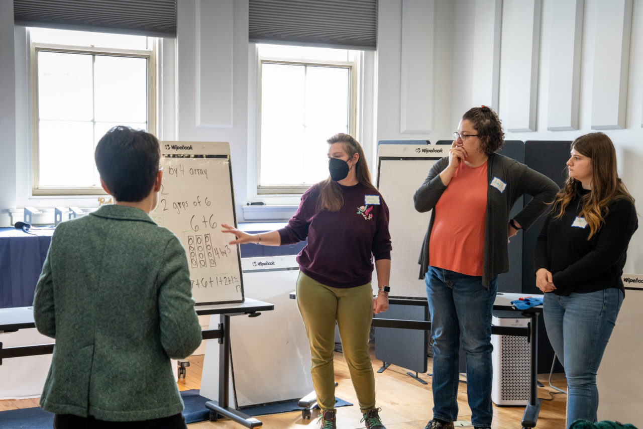 Educators discuss a math problem on a large notepad easel at an MMSA math workshop.