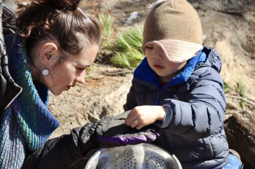 A child and an adult are shown inspecting dirt in an outdoor environment. Both look at something the child is putting in the adult's hand.