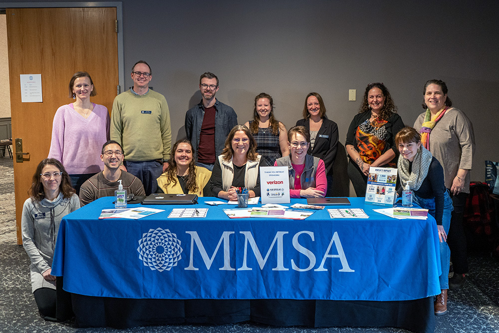 MMSA staff are shown in two rows behind a table with an MMSA-emblazoned tablecloth.