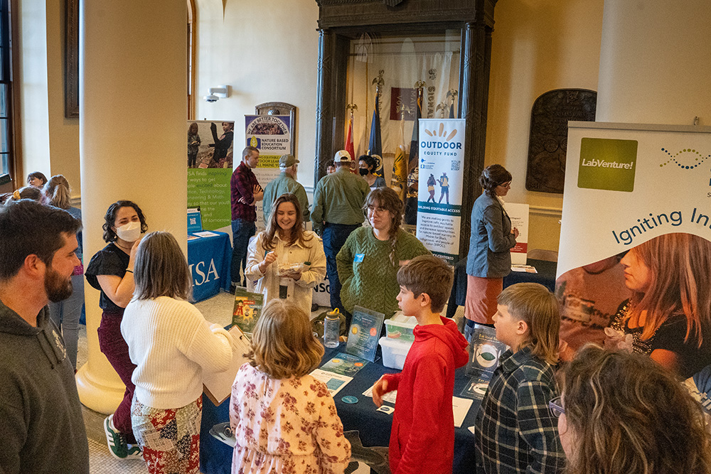 A mix of children and adults can be seen mingling in the State House in Augusta. Tables and signage for various organizations are set up.