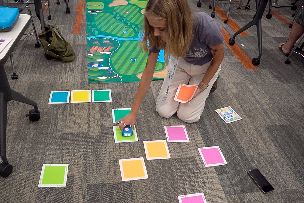 An educator kneels on the floor of a conference room working with a robot and colorful cards that the robot interacts with.