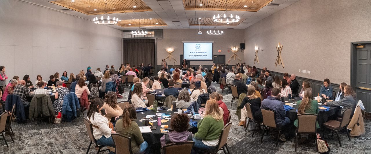 Participants are shown in a large conference room, with a projector screen at the front of the room displaying "STEM PD Retreat 2025."