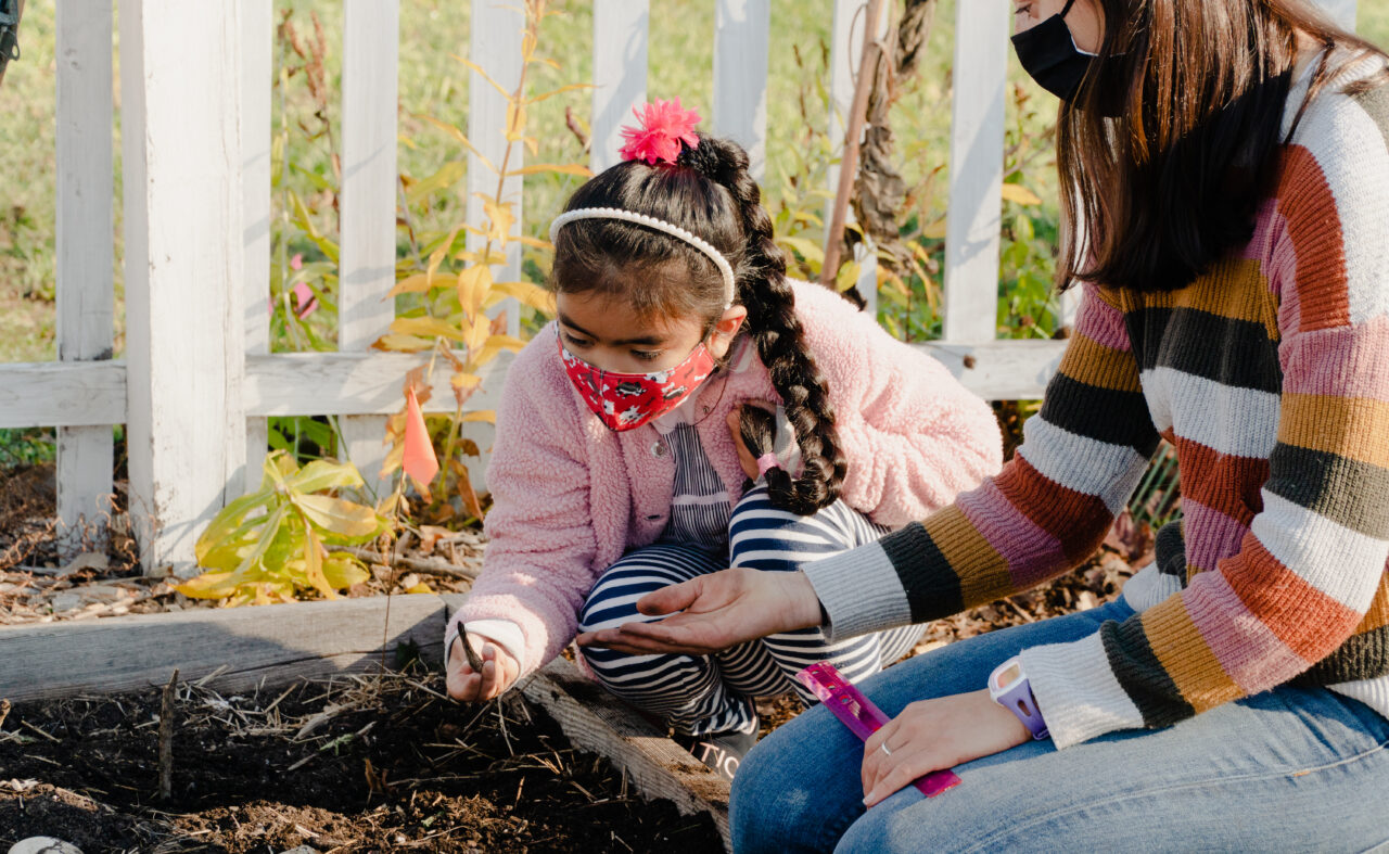 A child and an adult wearing medical masks look at plants in a garden.