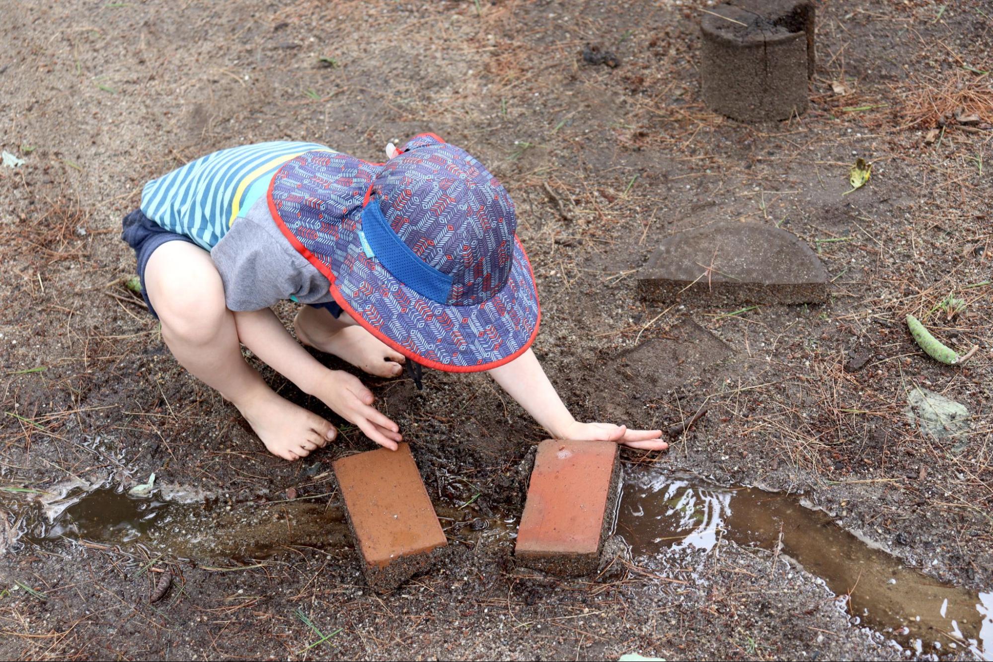 A child wearing a broad-brimmed rain hat arranges bricks on the ground in the mud.