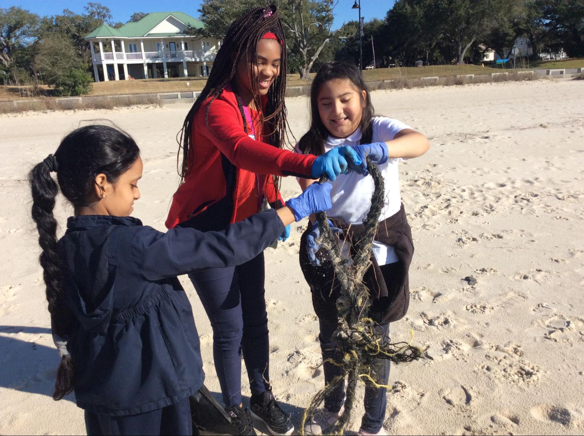 Three kids wearing plastic gloves pick up an object that washed up on the beach they are standing on.