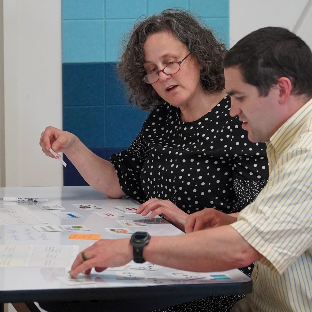 Educators and a facilitator discuss a post-sized white board propped on a table in a brightly lit conference room during a mathematics professional development workshop led by MMSA.