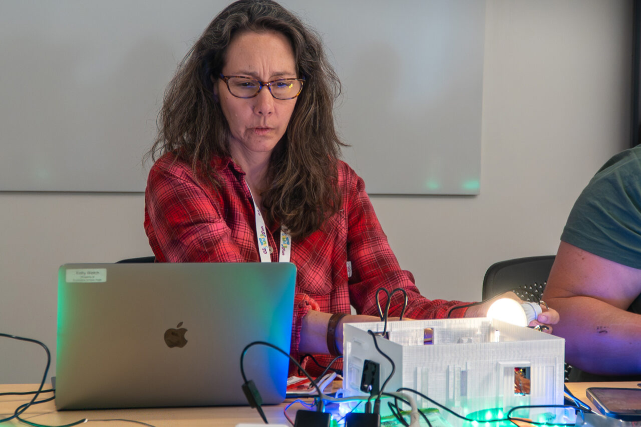 An educator holding a lightbulb works with a smart greenhouse, which is brightly lit, and a laptop computer.