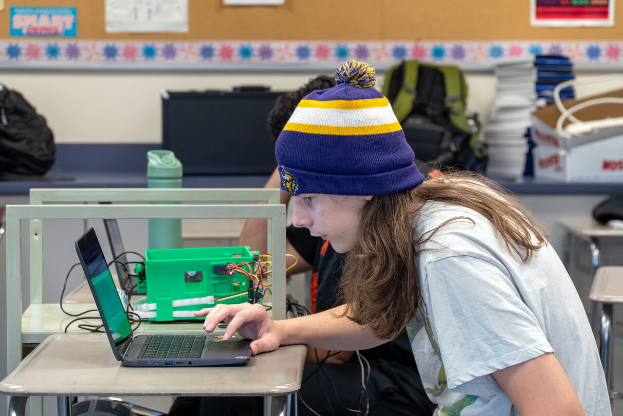 A student sits at a desk in a classroom while working on programming a smart greenhouse using their laptop.