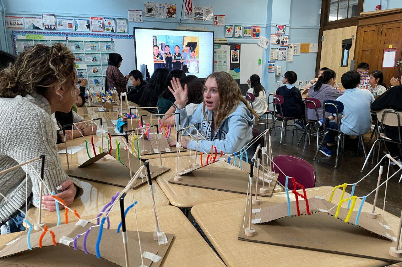 Kids at PS180 in Manhattan discuss an activity across school desks pushed together to form a long table. Several cardboard-and-string bridges are sitting on the table. Another set of students sit in the background, and an educator at the front of the room stands in front of a projector screen showing students holding the model bridges.