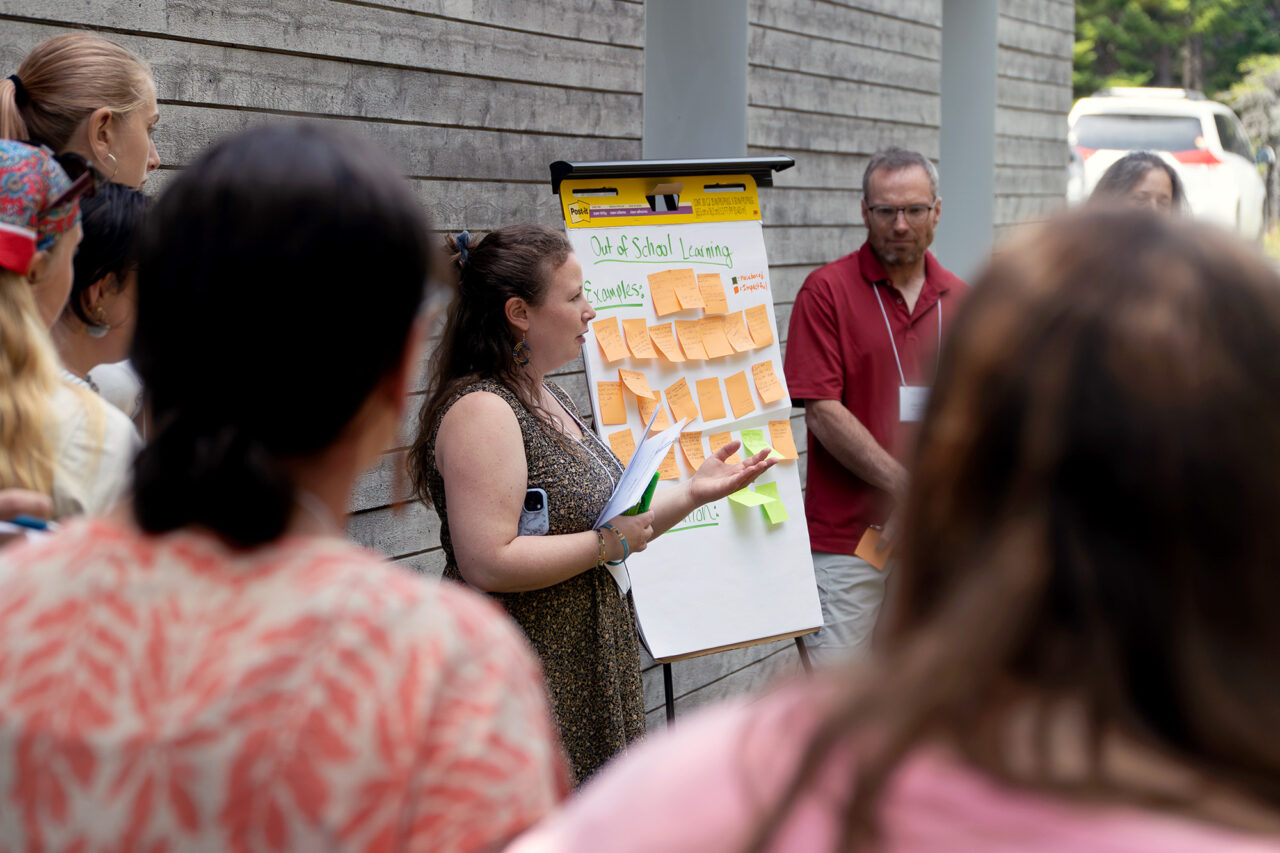 An MMSA staff person presents outside in front of a poster board on an easel and with sticky notes applied to it. A group is circled around the presenter and the poster board.