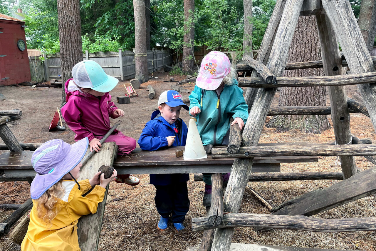 Young children play with different objects, including a small traffic cone and a stick, outside around a wooden play structure.