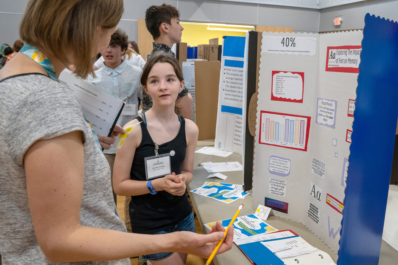A student appears to be listening to a question from an evaluator about their science fair project, which can be partially seen represented by a trifold on a table in the middleground.