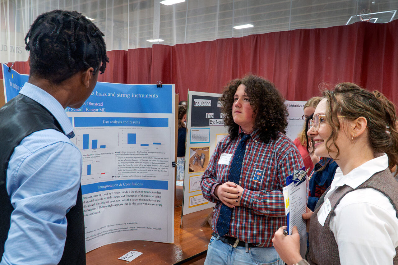 Participants in the middle school science fair discuss a poster about a project analyzing musical instrument frequencies.