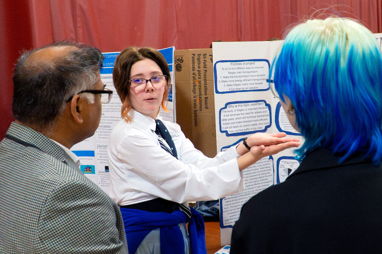 A student presents a project trifold to an evaluator and another student participant in the middle school science and engineering fair.