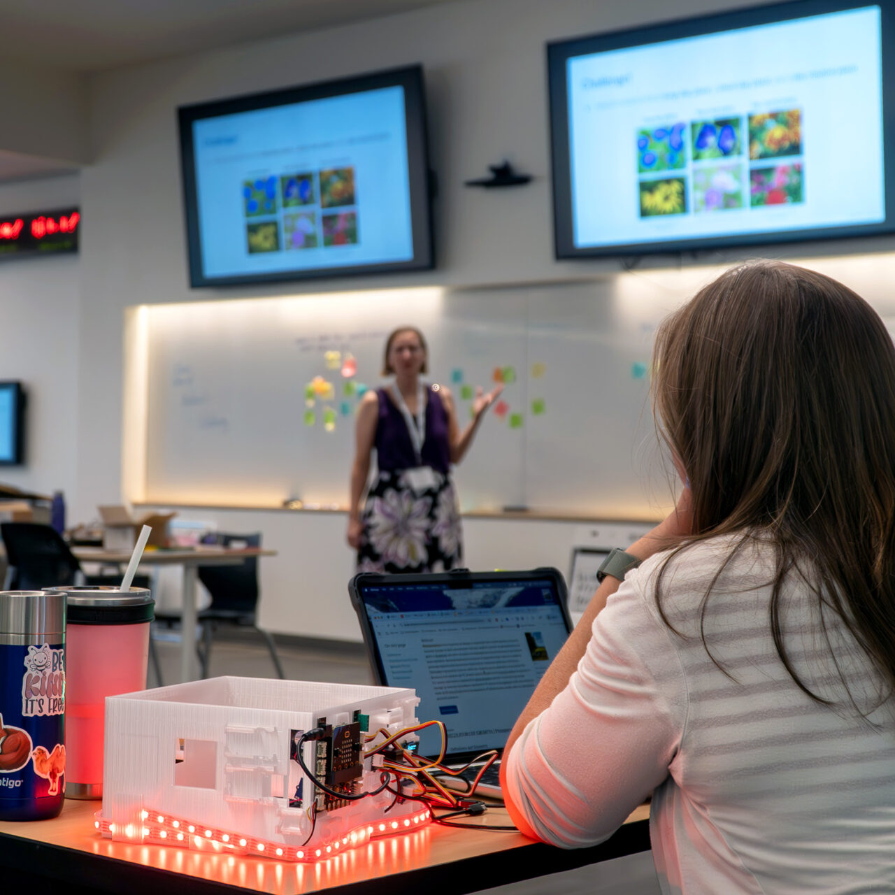 A participant is shown in the foreground sitting next to a smart greenhouse, which is lit up; in the background is a presentation being given by a facilitator at the CS Summer of Fun in summer of 2025.
