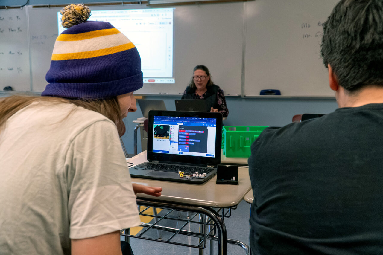 Students in a classroom work on a laptop showing block coding application that controls the small computer that is connected to sensors and lights inside the 3D-printed greenhouse, also on the desk. In the background, a teacher presents on the coding application.