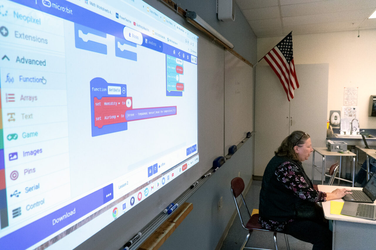 A teacher sits at a computer at a desk, behind which is a projection of some block code.