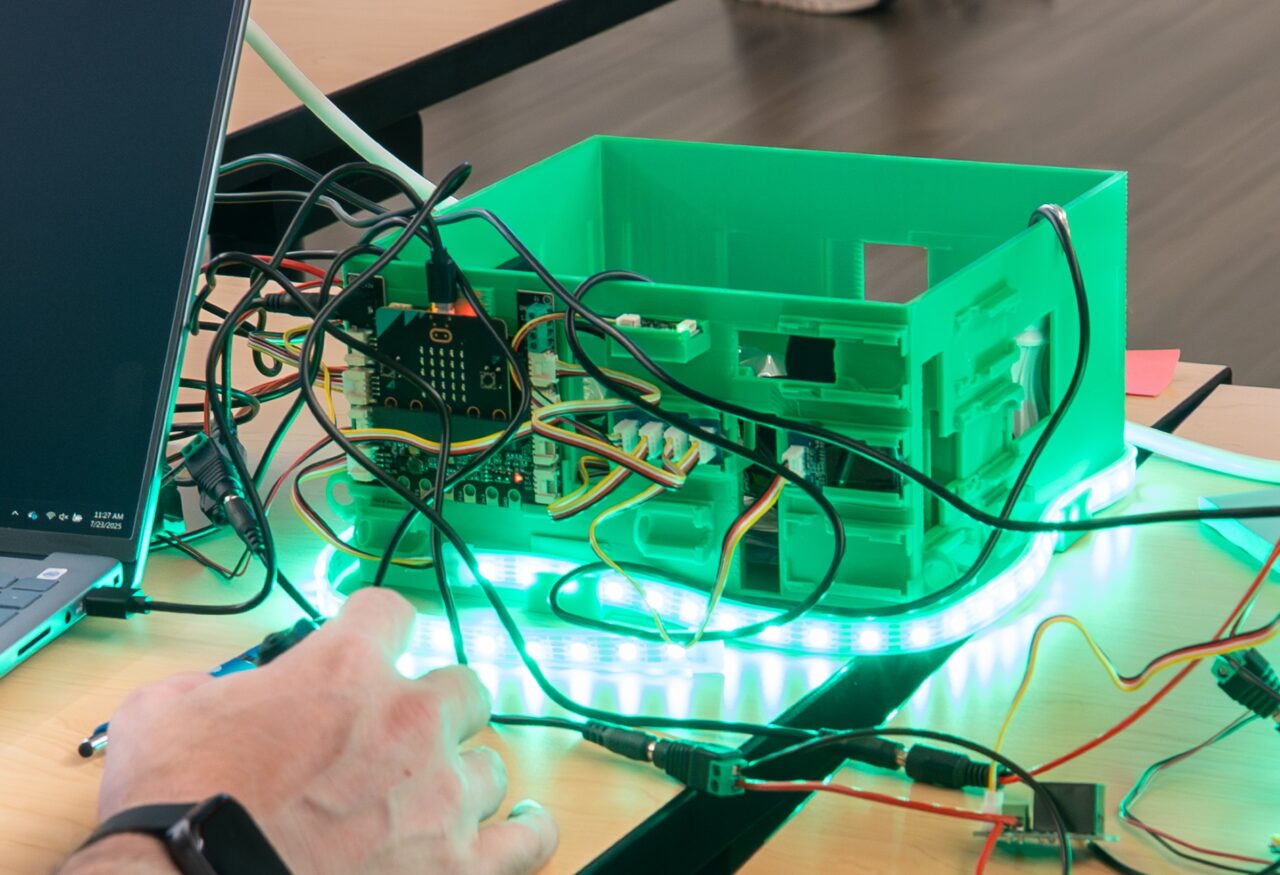 A green 3D-printed box sits on a table; there are a lot of cable connecting it to illuminated LED lights and a laptop. Someone's hand is interacting with a component at the edge of the frame.