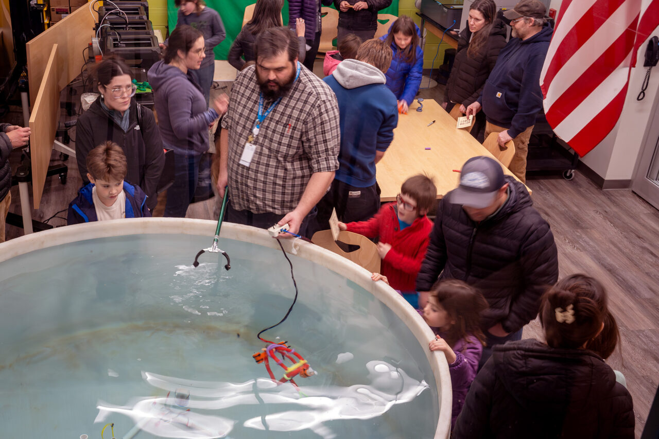 A pool is shown in the foreground and people around it are variously watching a demonstration of a robot that goes into the pool or working on robots in the background.