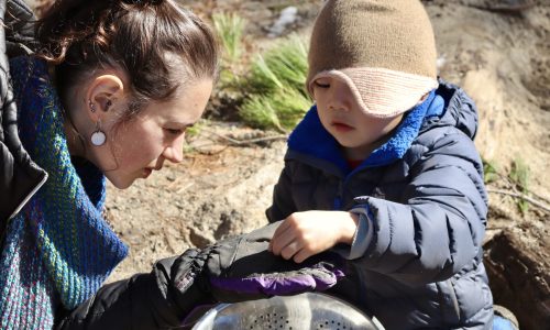 A child and an adult are shown inspecting dirt in an outdoor environment. Both look at something the child is putting in the adult's hand.