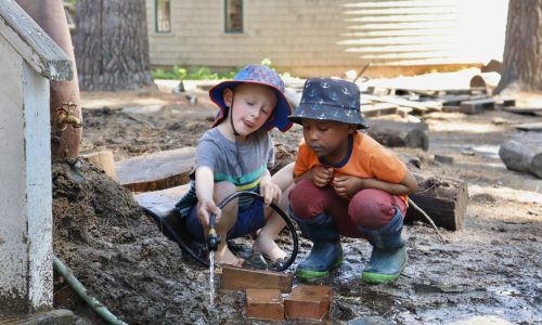 Two children wearing bucket hats crouch down outside next to a puddle. Both children stare intently at a stream of water one child is positioning over the mud using a garden hose.